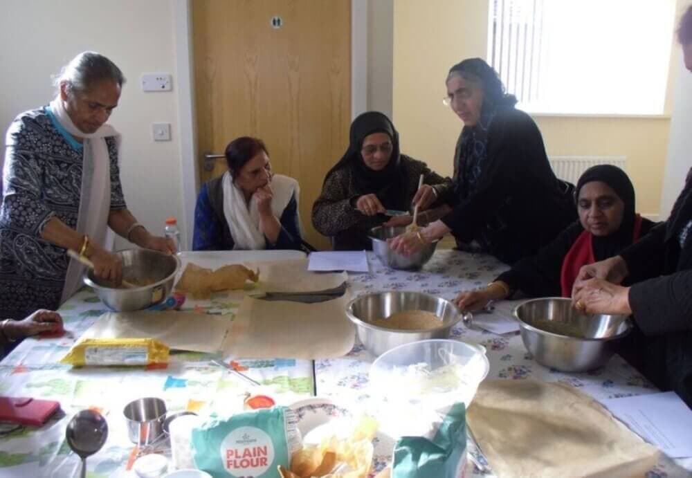A group of women gathered around a table preparing food, with ingredients and mixing bowls in front of them. - Home Instead