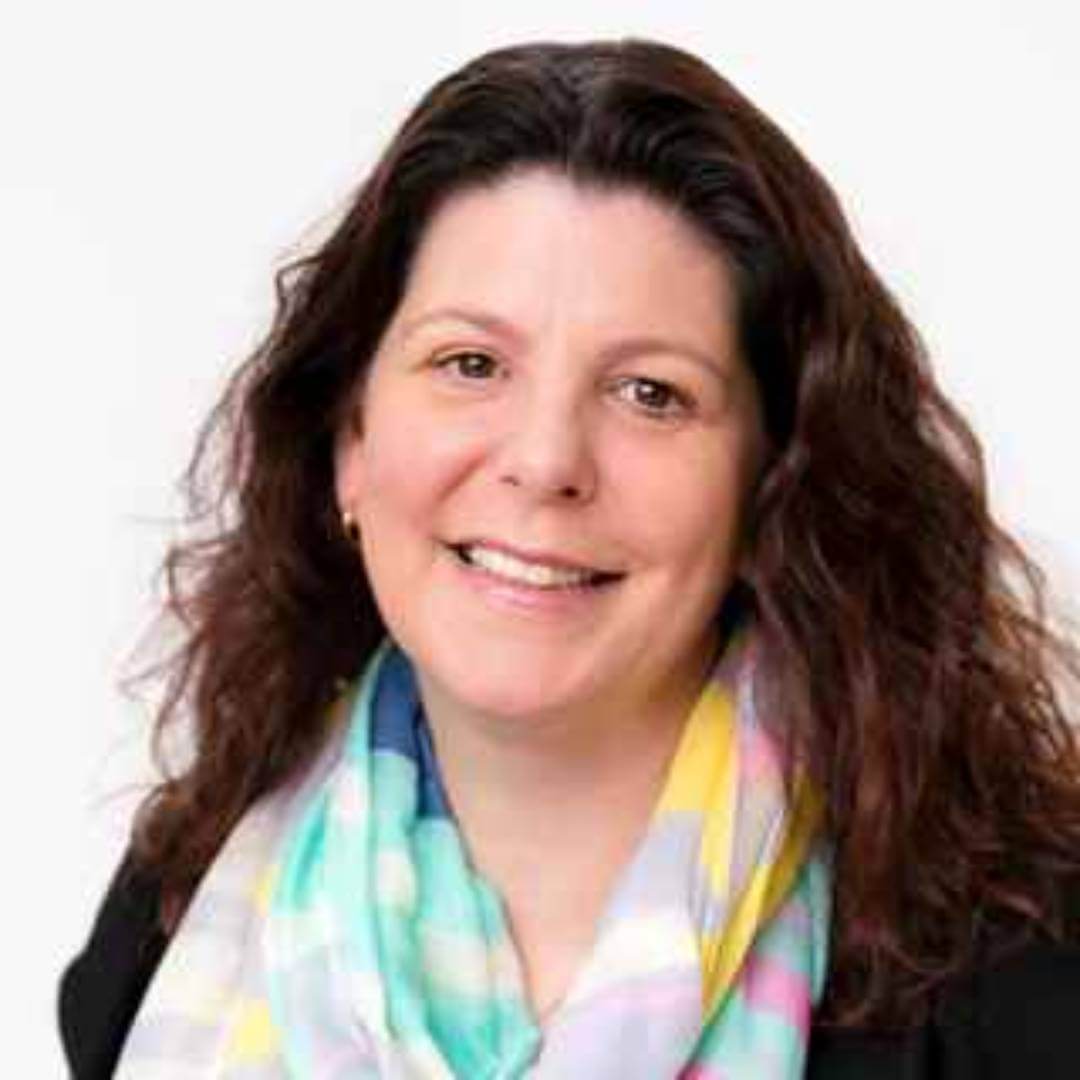 Smiling person with long brown hair, wearing a colorful scarf and black top, against a plain background. - Home Instead