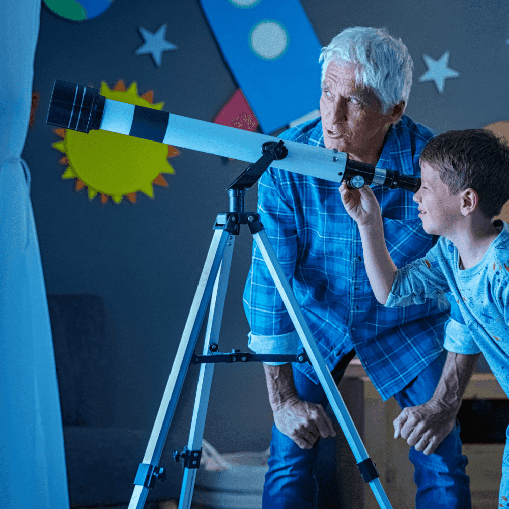 An elderly man and a young boy looking through a telescope indoors, with space-themed decorations in the background. - Home Instead