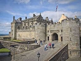 Historic stone castle with a flag on a pole and groups of tourists walking along the path and entryway. - Home Instead