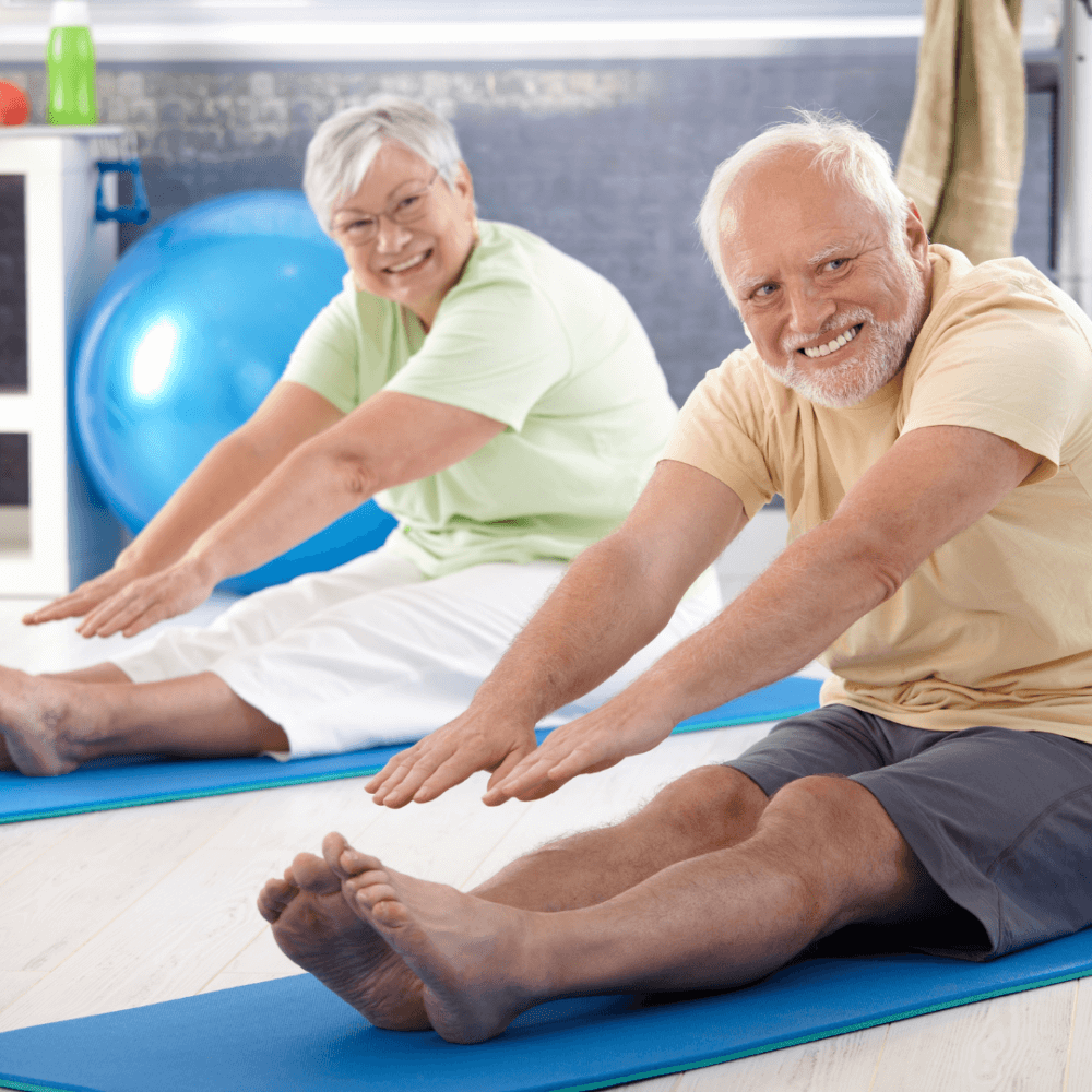 Two elderly individuals smiling and stretching on yoga mats indoors, with a blue exercise ball in the background. - Home Instead