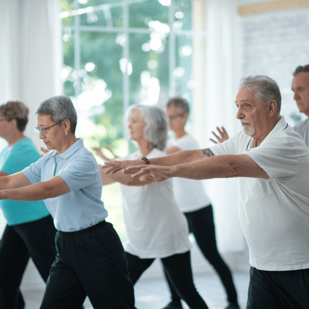 Older adults practicing tai chi in a bright, airy studio with large windows in the background. - Home Instead