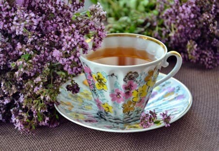 Floral teacup with tea on matching saucer surrounded by fresh purple flowers on a brown fabric background. - Home Instead