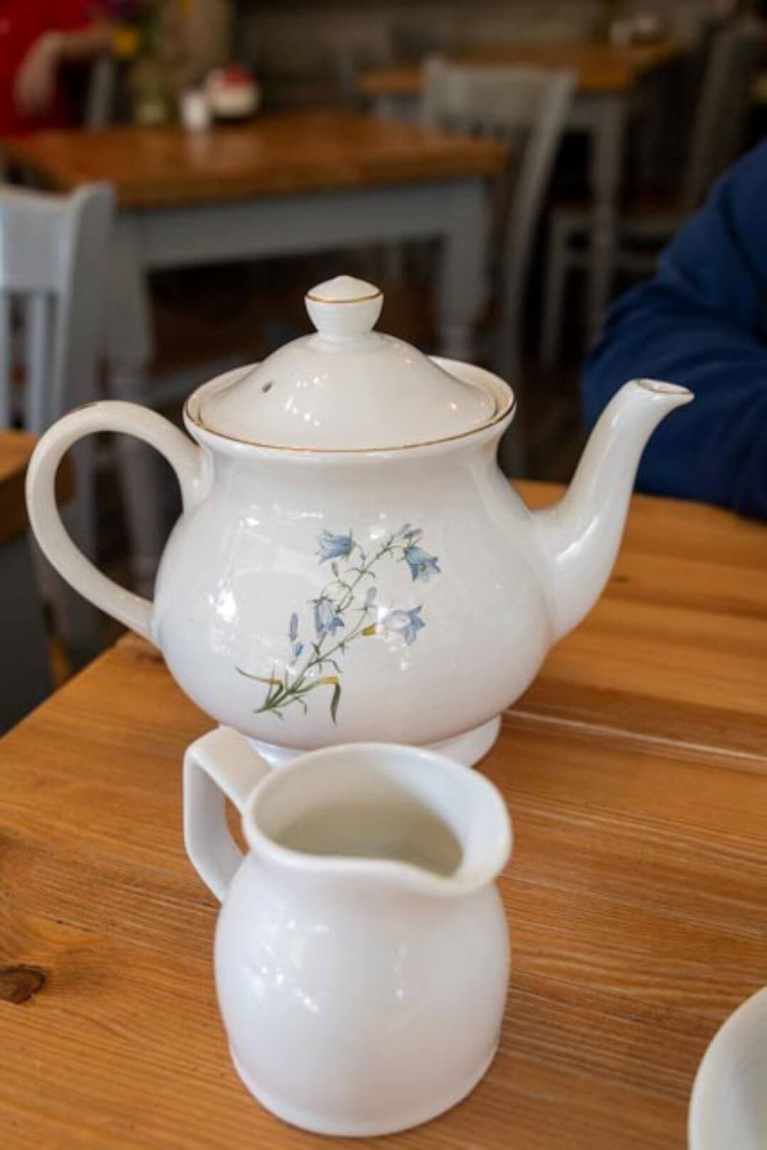 White teapot with blue floral design and matching creamer jug on a wooden table in a cozy cafe setting. - Home Instead