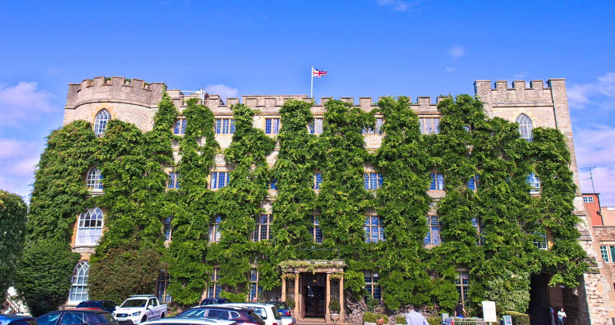 A historic stone building covered in ivy, flying a UK flag, with multiple cars parked in front under a clear blue sky. - Home Instead