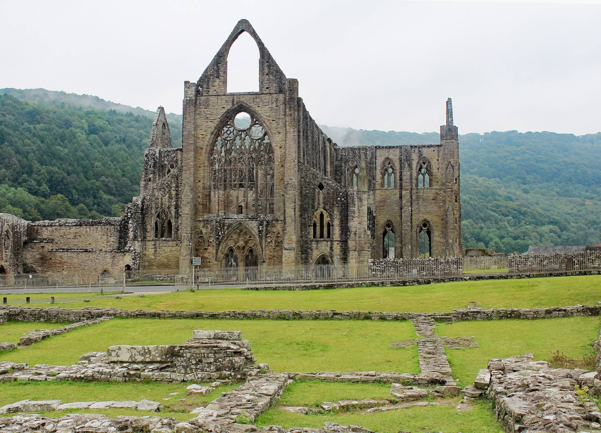 Ruins of a large stone abbey with arched windows on a grassy landscape, surrounded by forested hills. - Home Instead