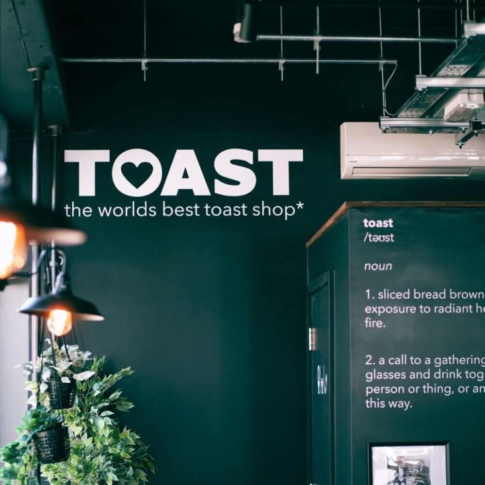 A wall with the sign "TOAST the world's best toast shop" and a large, playful dictionary definition of "toast. - Home Instead