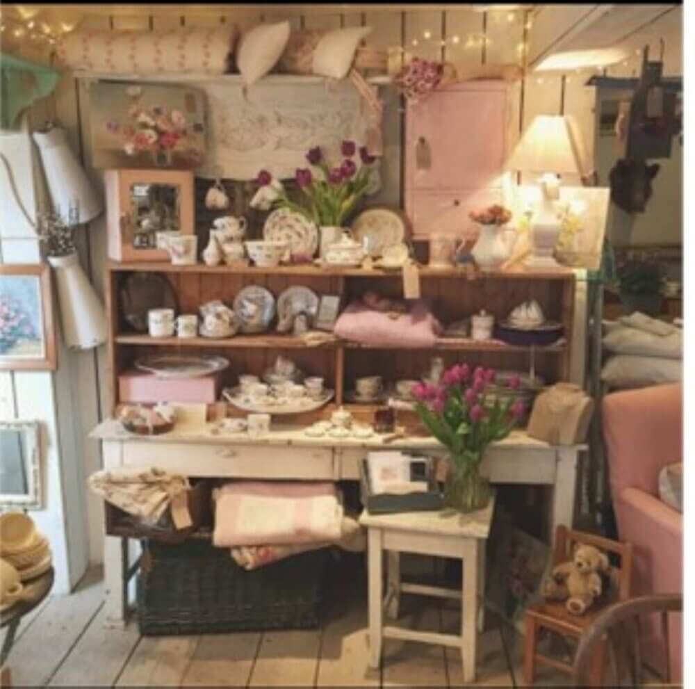 Shabby chic-style room with shelves displaying vintage teacups, plates, tulips, books, linens, and a teddy bear. - Home Instead