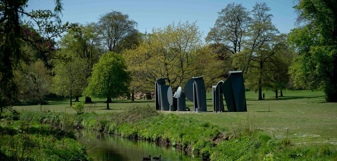 Outdoor sculpture installation in a green park with trees, a pond, and ducks swimming in the foreground. - Home Instead