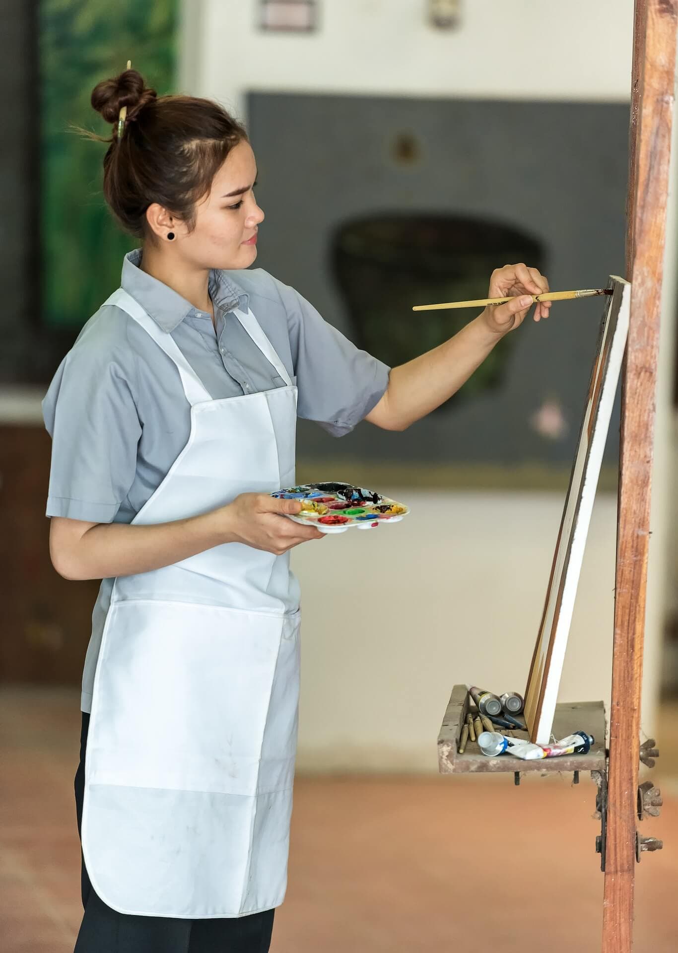 Woman painting on a canvas with a brush and palette in hand, wearing a gray shirt and white apron. - Home Instead