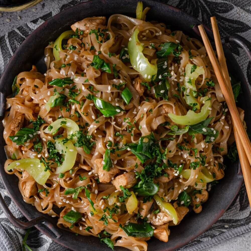 A bowl of chicken stir-fry noodles garnished with sesame seeds, green onions, and leafy greens, with chopsticks on the side. - Home Instead