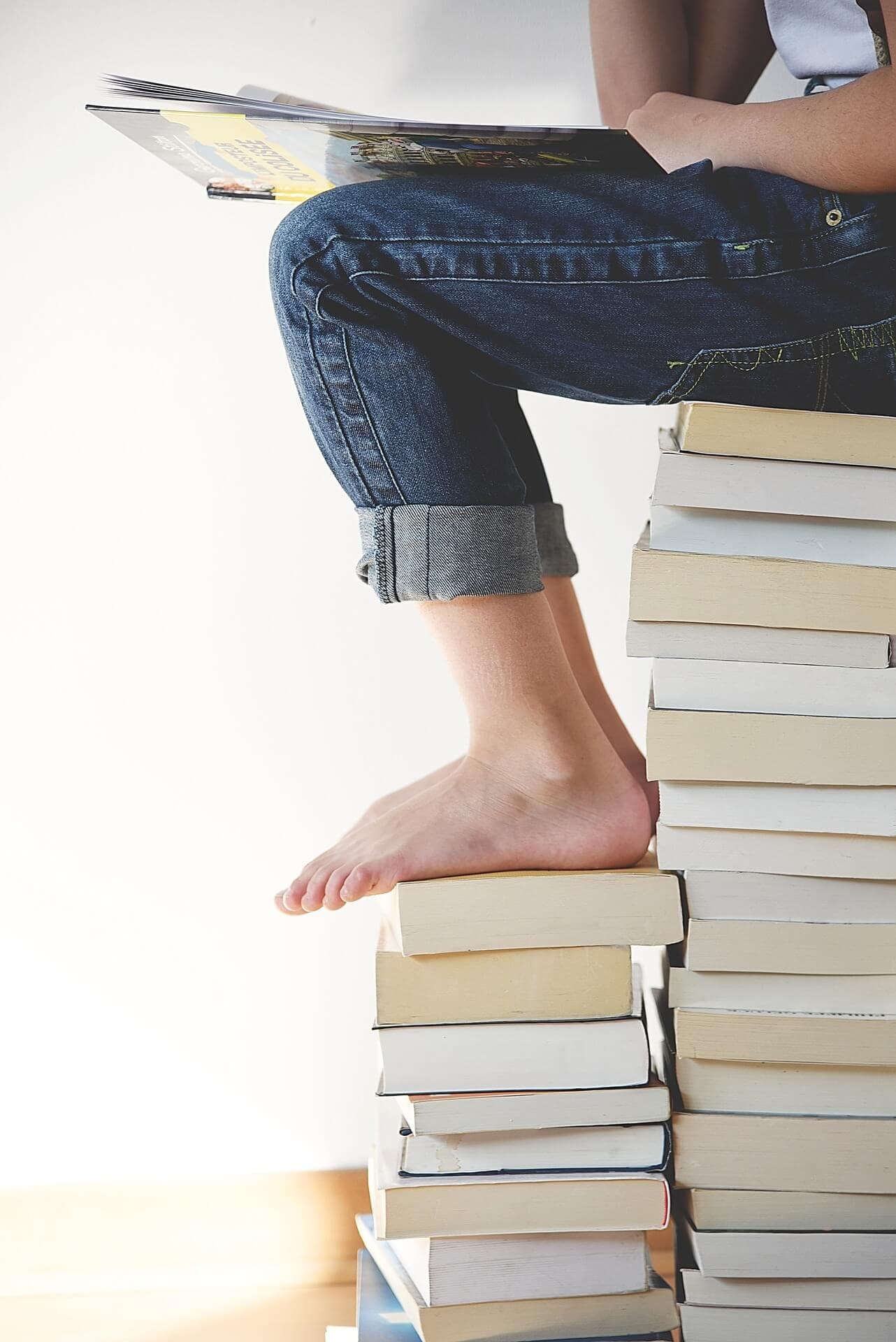 A person sitting on a tall stack of books, reading. They are barefoot and wearing rolled-up jeans. - Home Instead