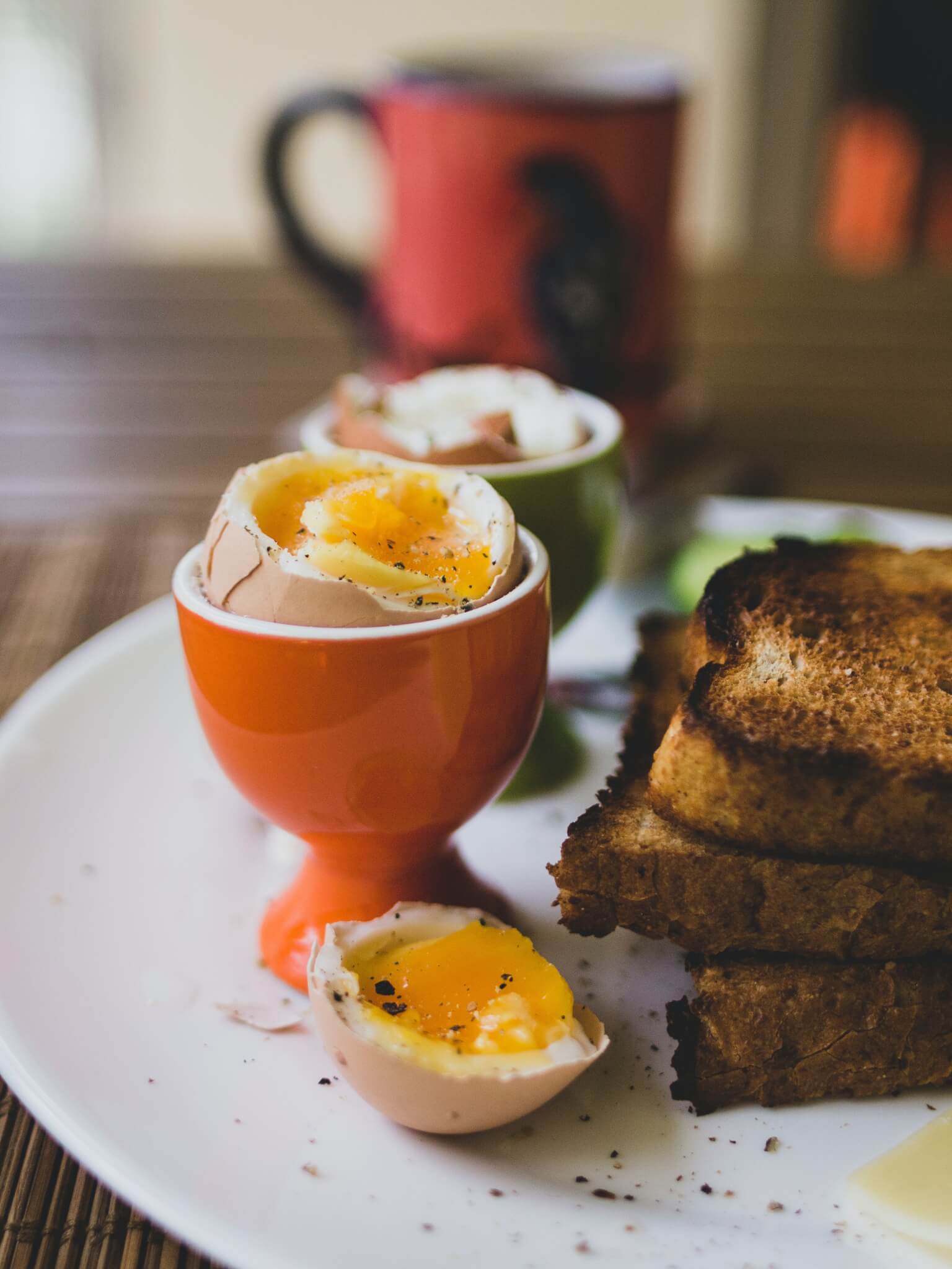 Soft-boiled eggs in egg cups with toast on a white plate. A mug in the background. - Home Instead