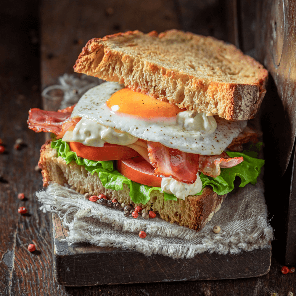 A sandwich with fried egg, bacon, lettuce, tomato, and mayonnaise between two slices of toasted bread on a rustic cloth. - Home Instead