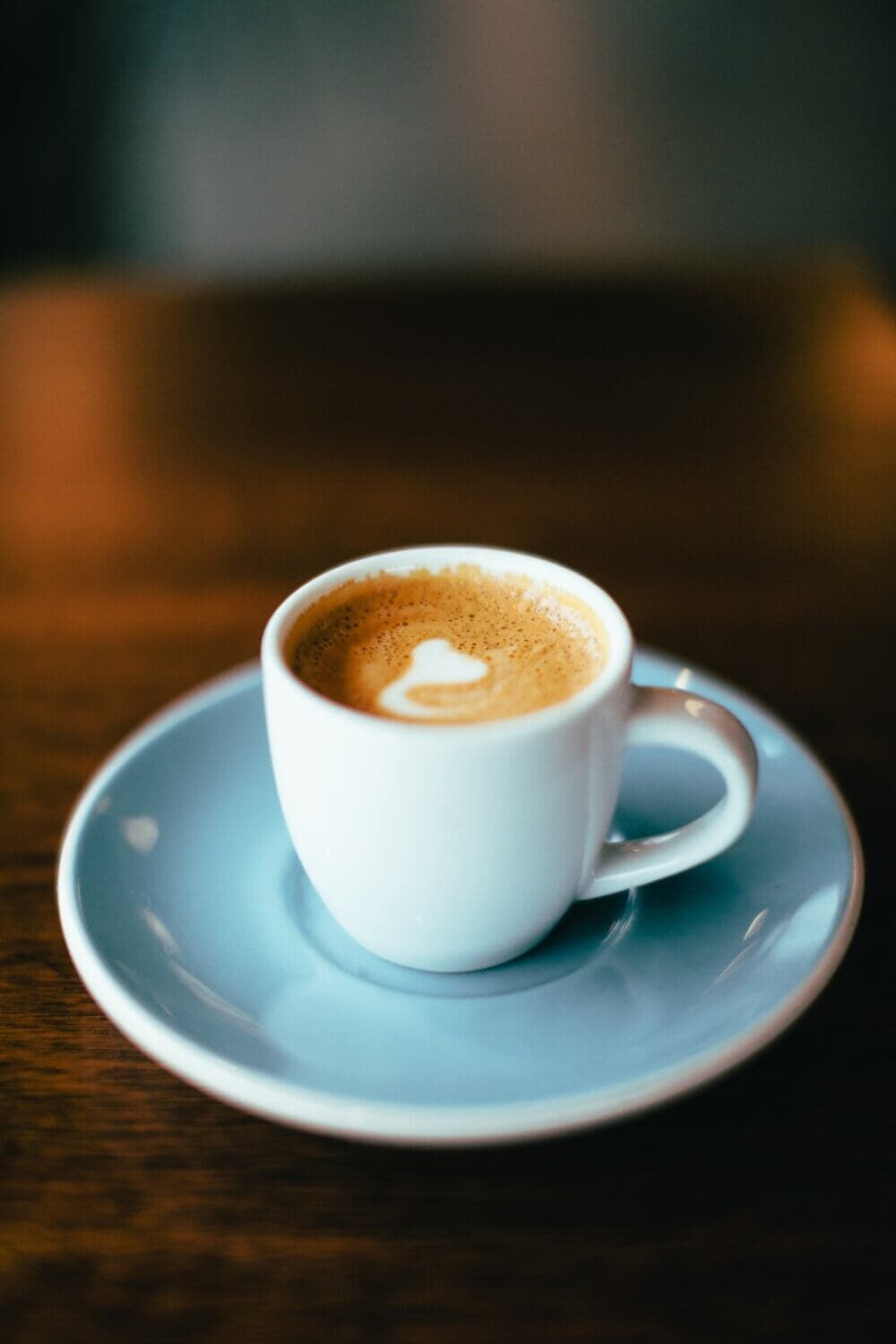 A cup of espresso with latte art in a white cup and saucer on a wooden table. - Home Instead