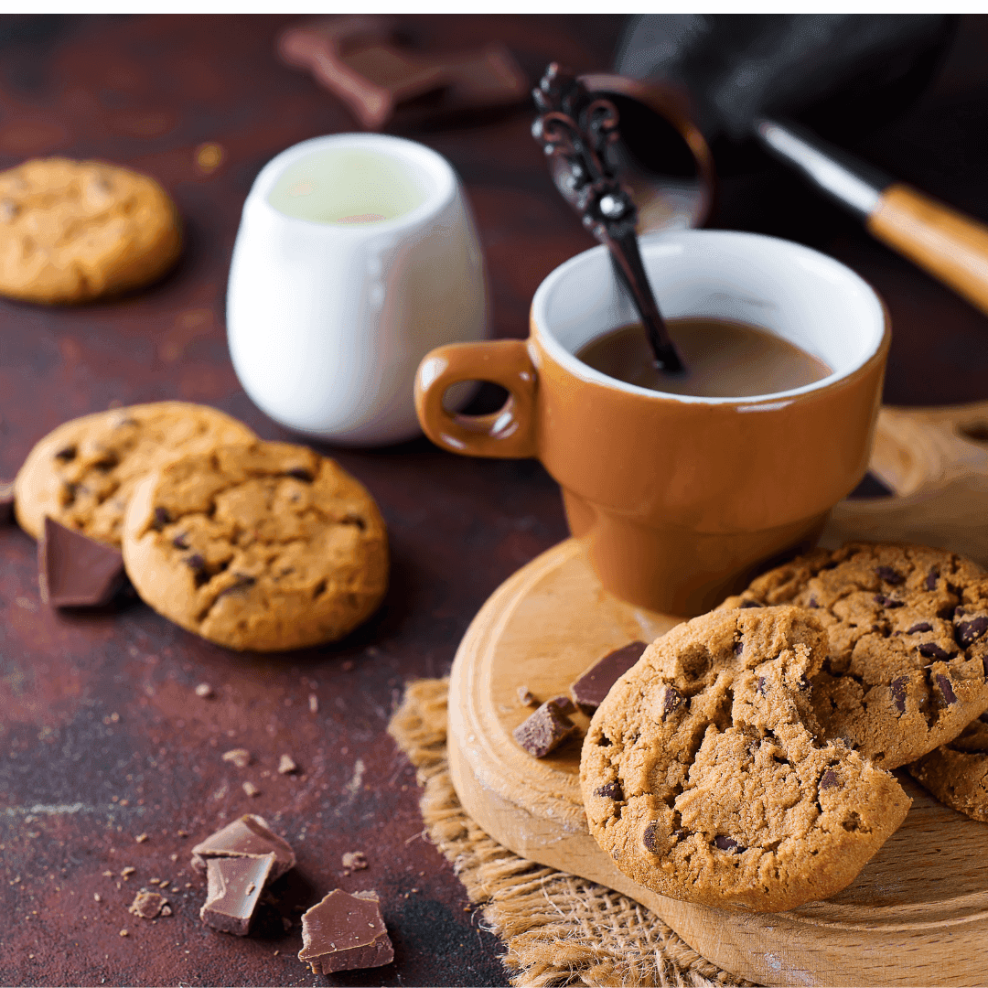 Chocolate chip cookies with a brown coffee cup, spoon, milk jug, and chocolate pieces on a rustic surface. - Home Instead