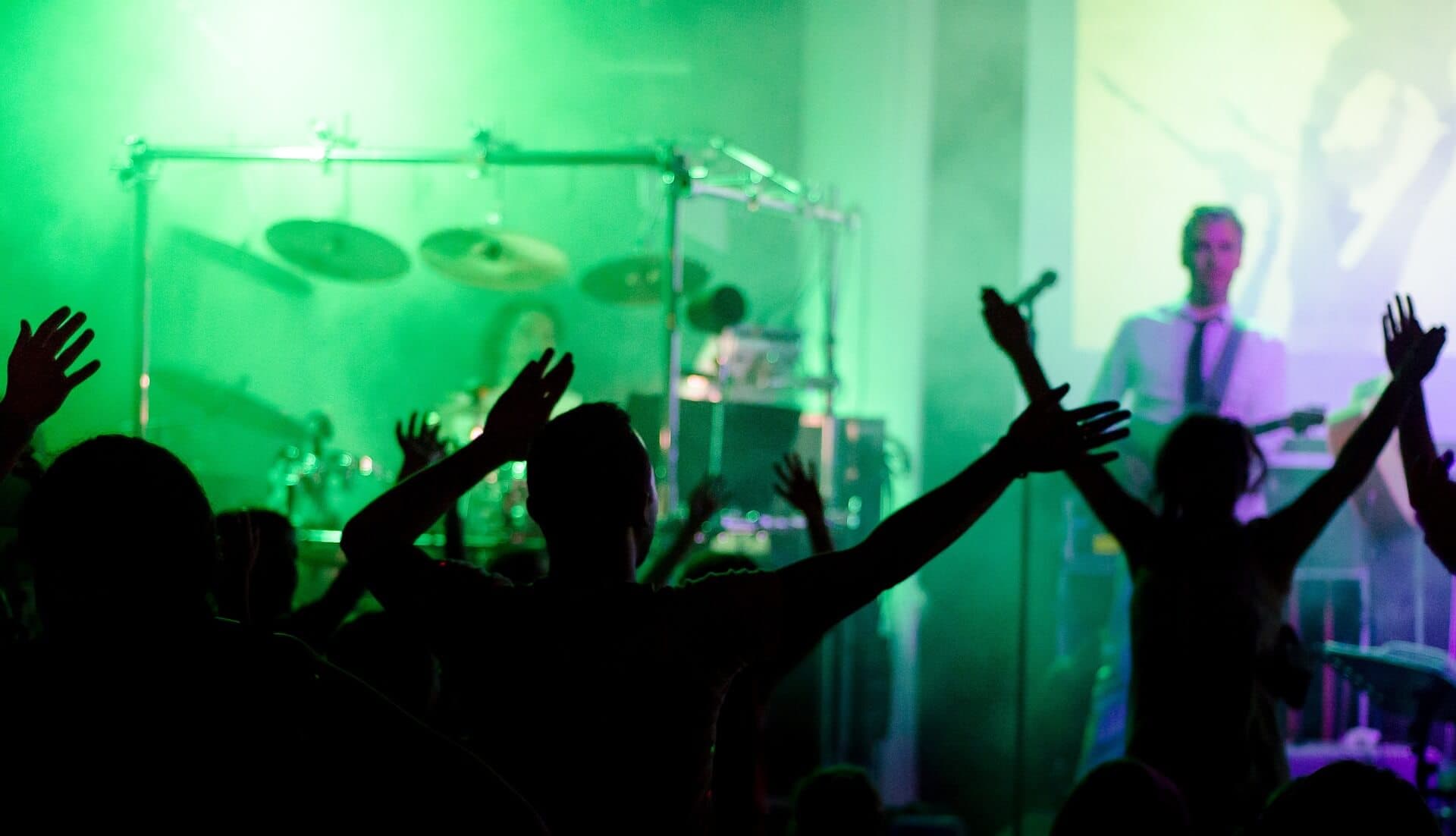 Silhouetted audience with raised hands at a concert, green lighting and musicians performing in the background. - Home Instead