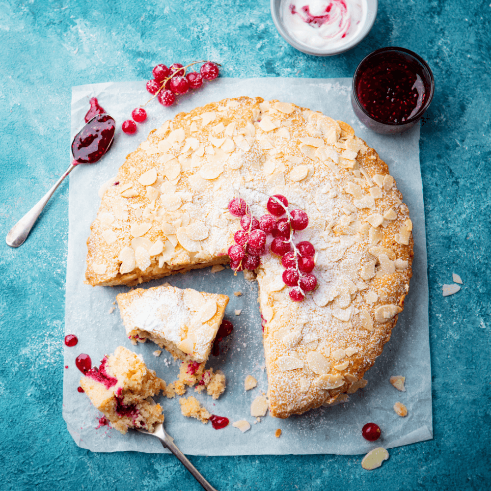 A round almond cake with powdered sugar and red currants on top, placed on parchment paper with jam and cream nearby. - Home Instead