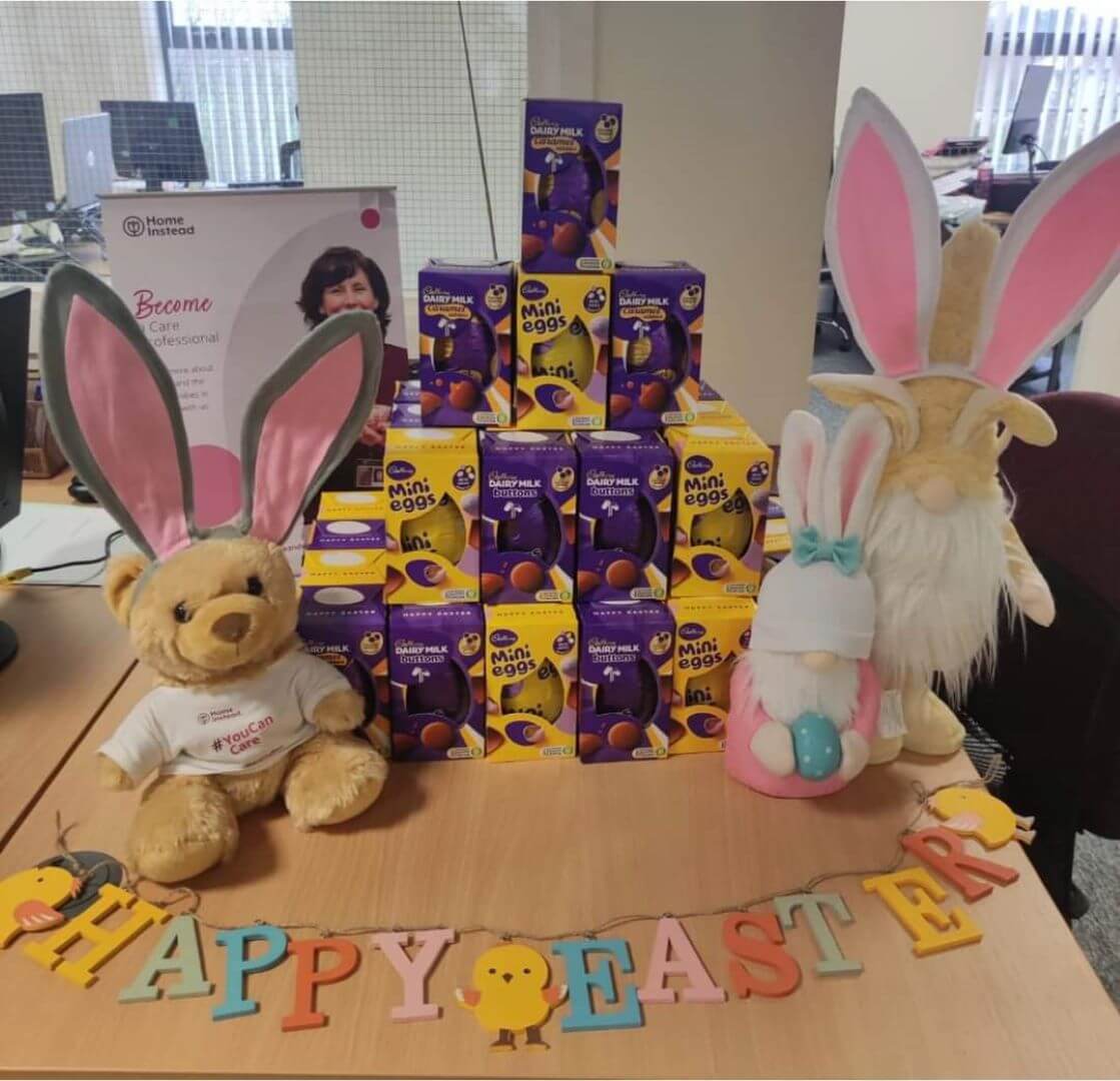Plush toys with bunny ears and Easter decorations arranged around stacks of Cadbury Mini Eggs boxes. "Happy Easter" banner below. - Home Instead