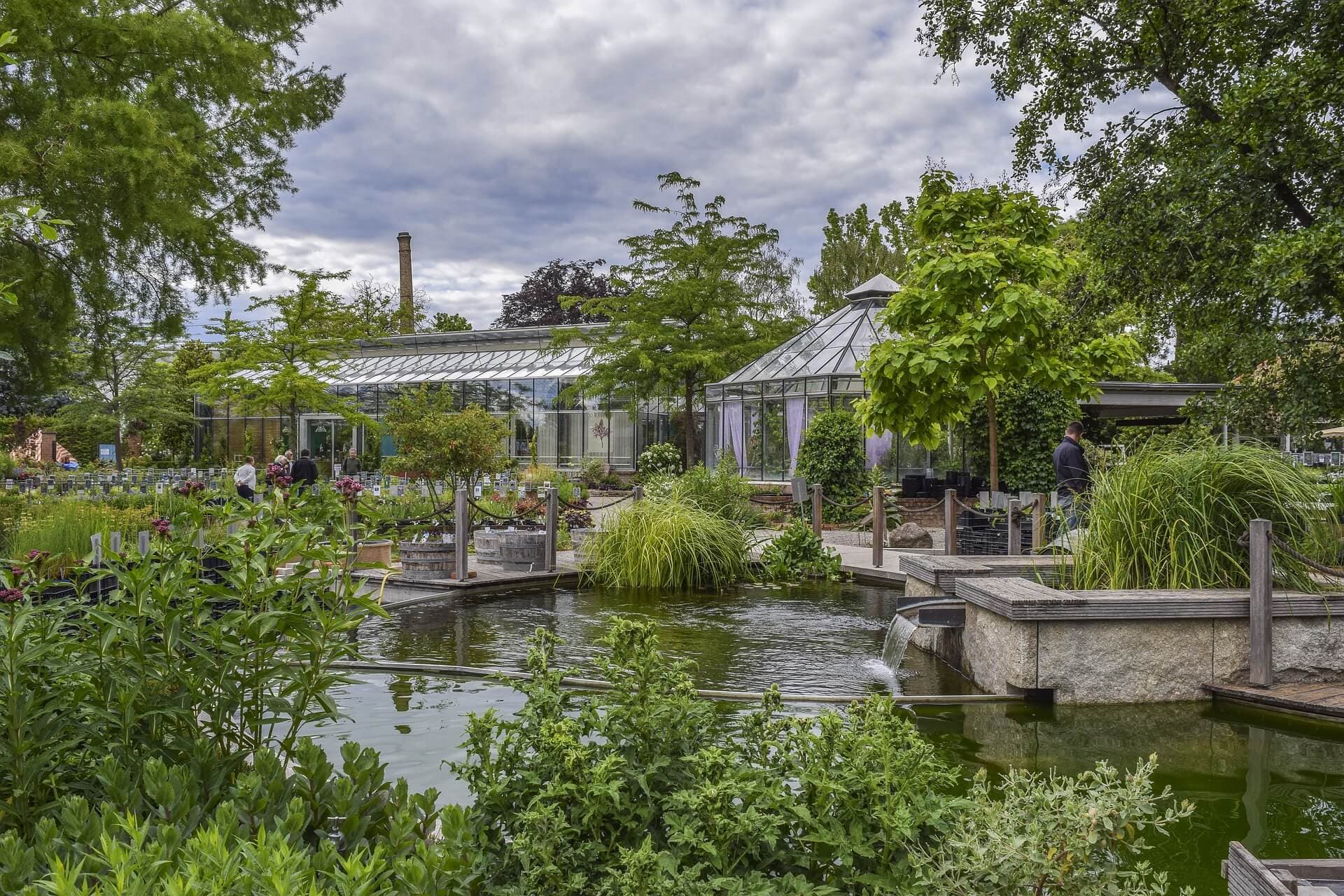 Lush botanical garden with a pond, greenery, and glass greenhouses under a cloudy sky. People are walking in the background. - Home Instead