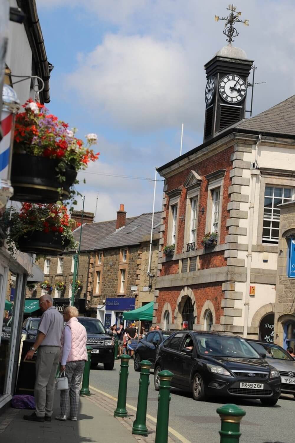 A busy town street with people, cars, a building with a clock tower, and a shop with hanging flower baskets. - Home Instead