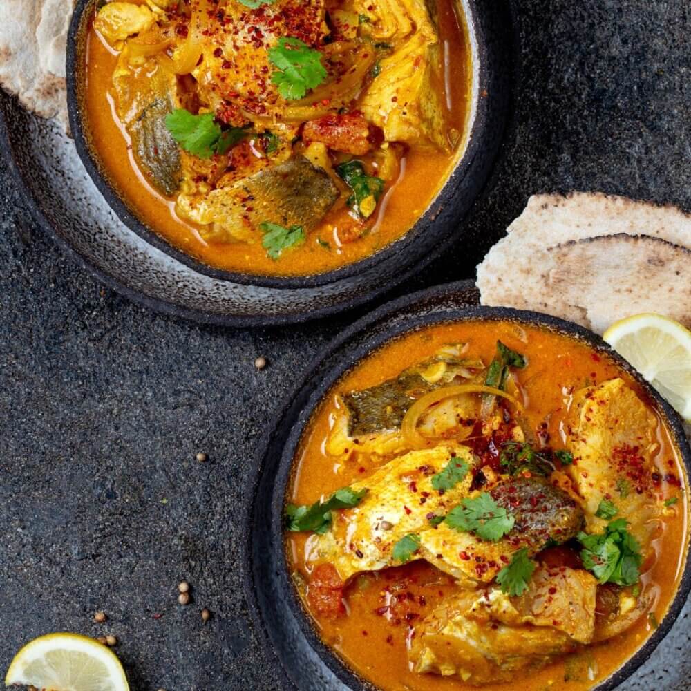 Two bowls of fish curry garnished with herbs, served with lemon slices and flatbread on a dark textured surface. - Home Instead
