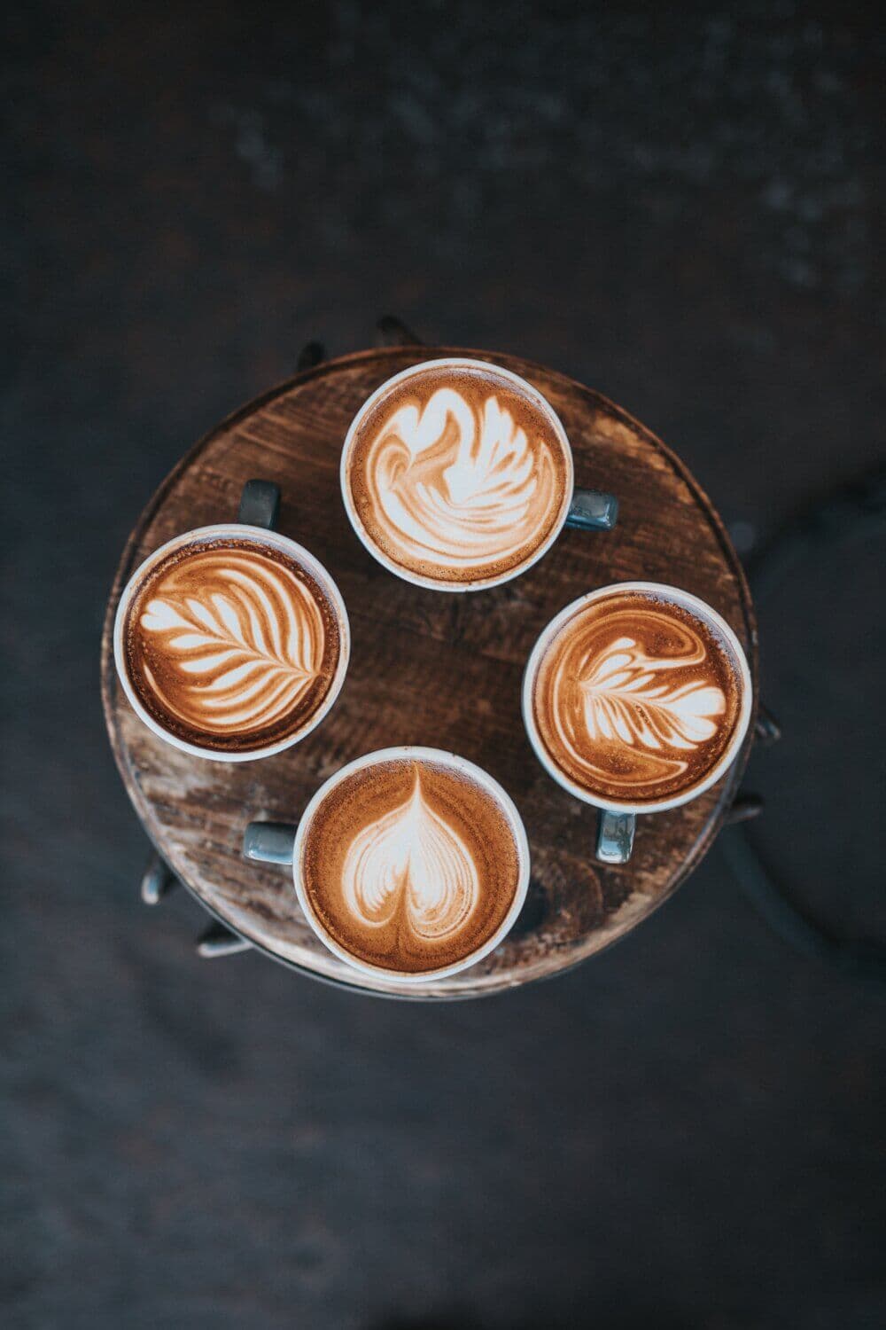 Top view of four mugs of latte with different latte art designs, placed on a round wooden surface. - Home Instead