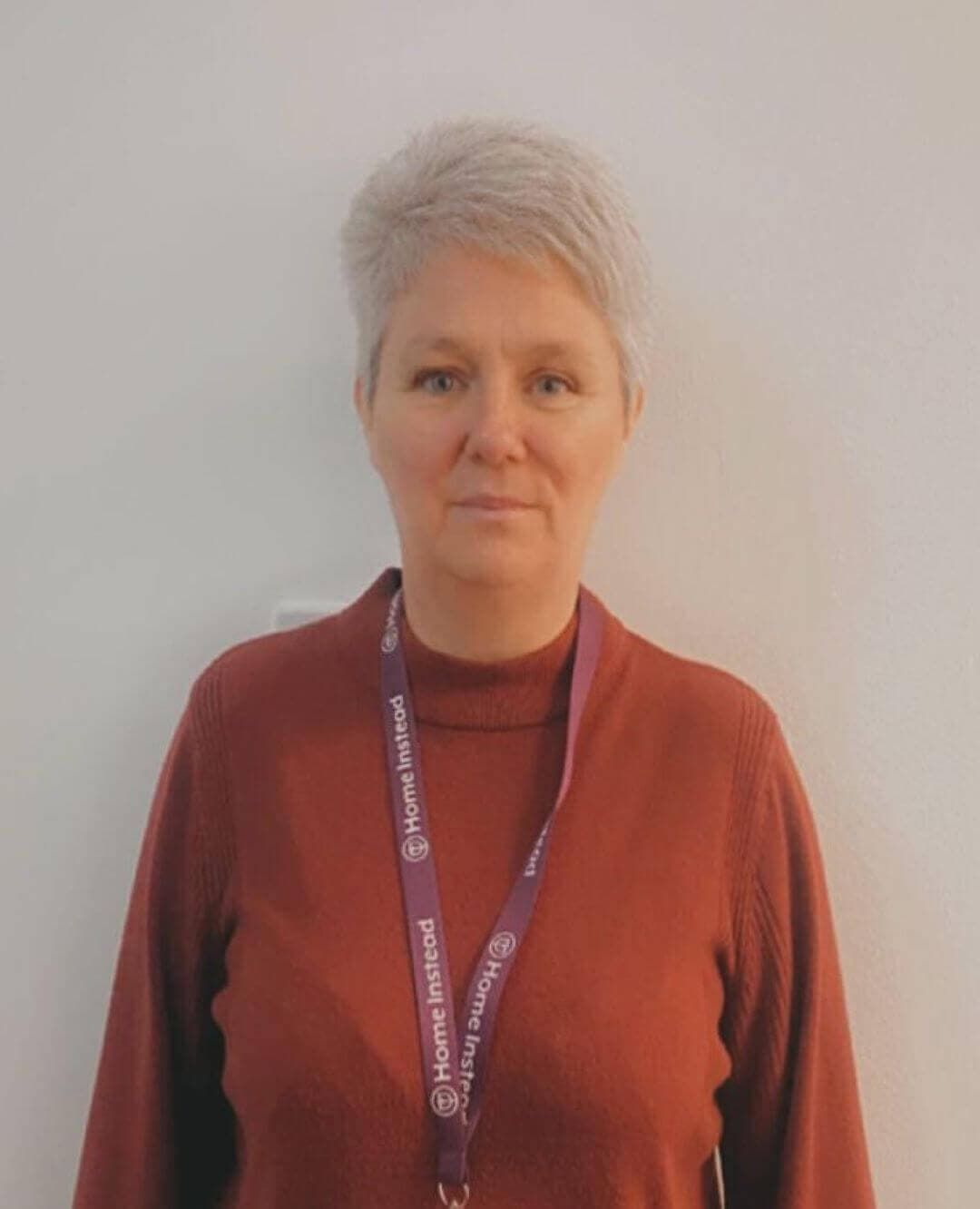 Person wearing a red sweater and a lanyard with “Home Instead” logo, standing against a plain white wall. - Home Instead