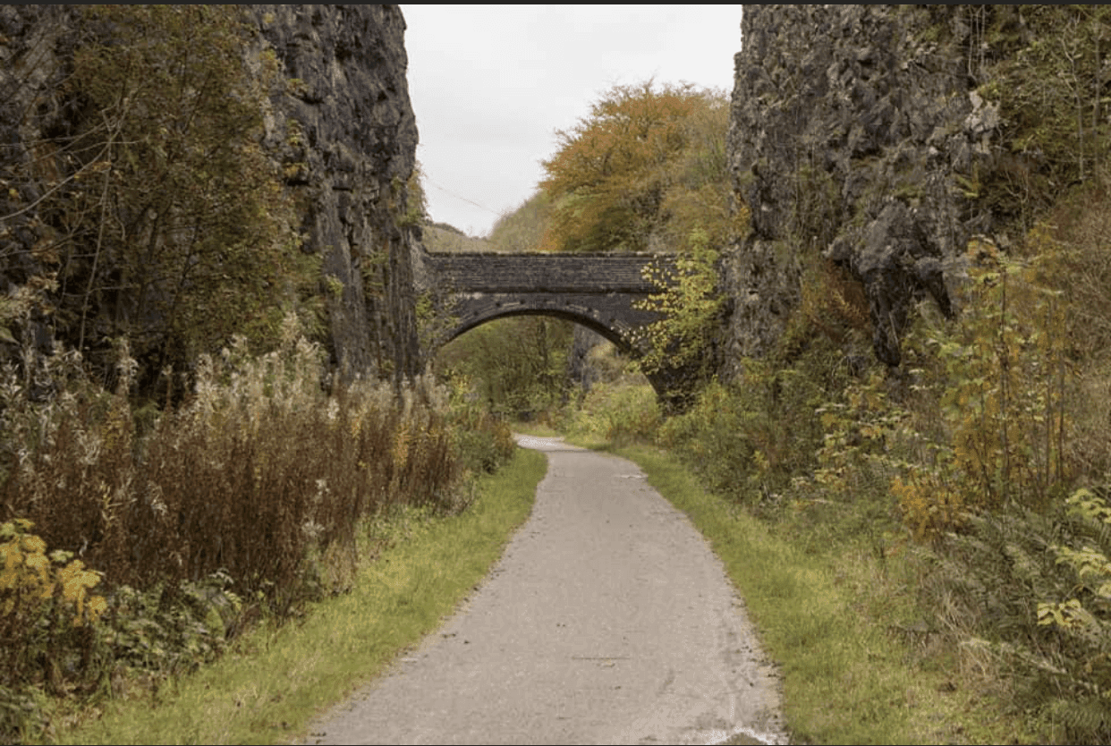 A narrow path leads under a stone bridge surrounded by tall rock cliffs and autumn foliage. - Home Instead