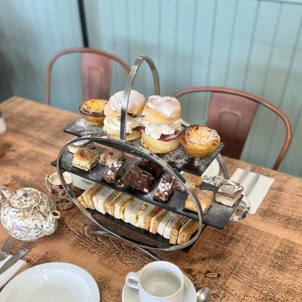 Afternoon tea set with a tiered stand of sandwiches, pastries, and desserts on a wooden table with teacups and teapot. - Home Instead