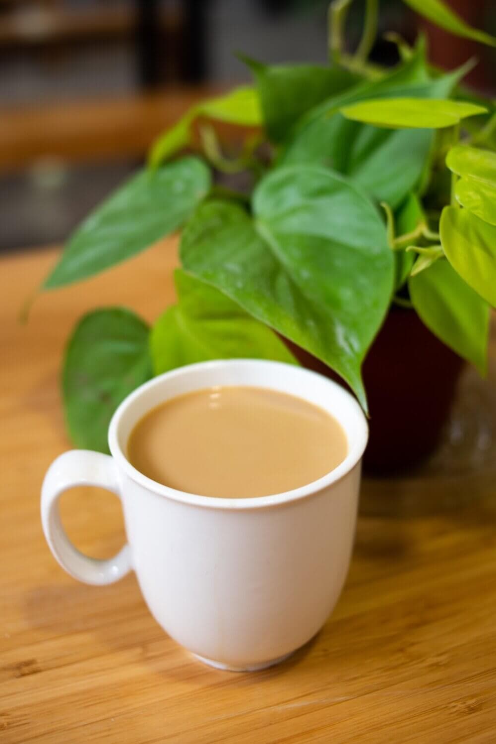 A white mug filled with coffee sits on a wooden table next to a green potted plant with heart-shaped leaves. - Home Instead