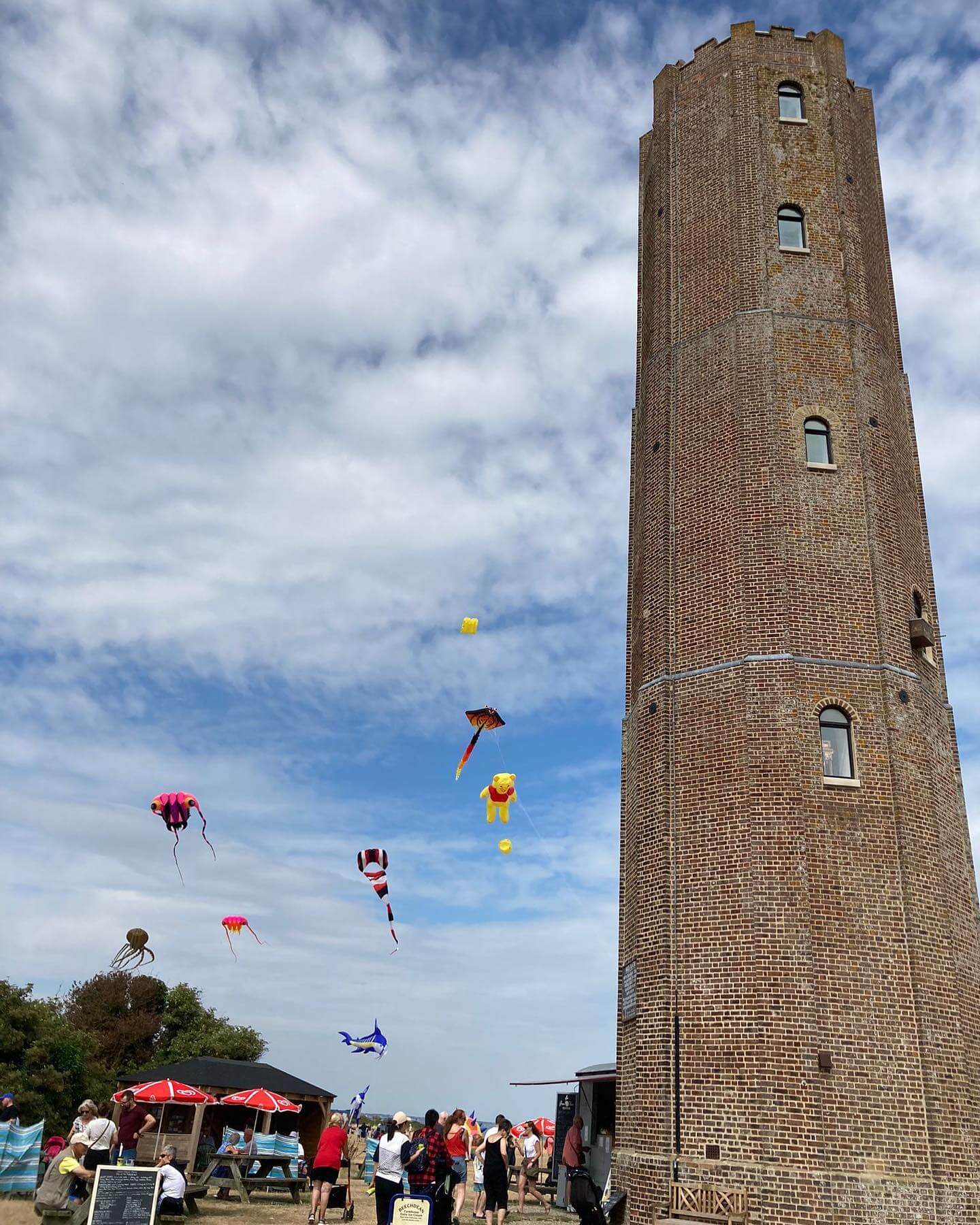 Tall brick tower with people flying colorful kites nearby under a blue sky with clouds. - Home Instead