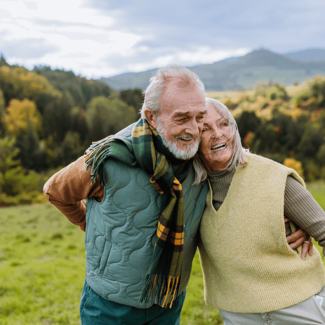 An elderly couple smiling and embracing outdoors in a scenic, mountainous landscape with autumn foliage. - Home Instead
