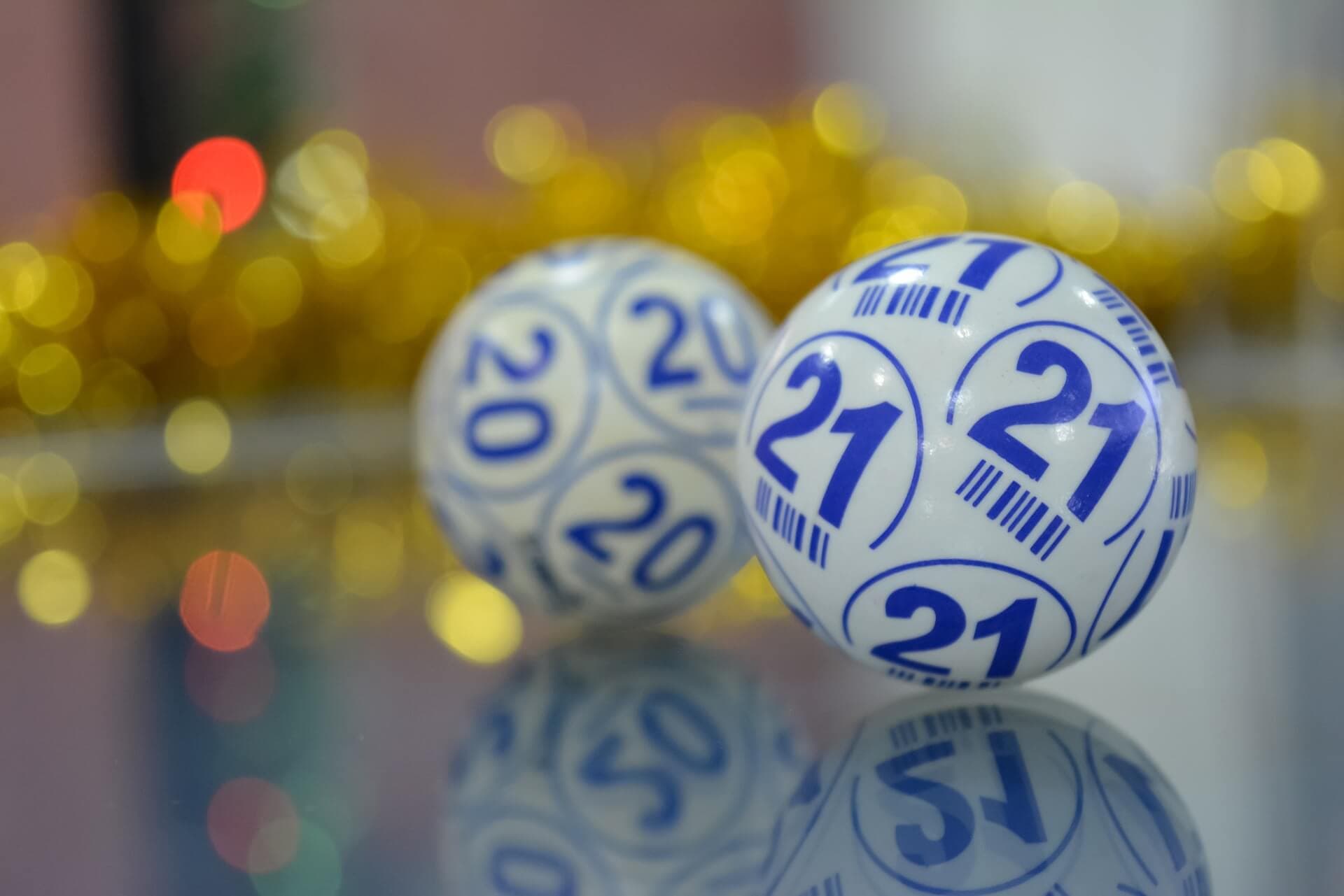 Close-up of lotto balls numbered 20 and 21 with a blurred background decorated with gold tinsel and colorful lights. - Home Instead