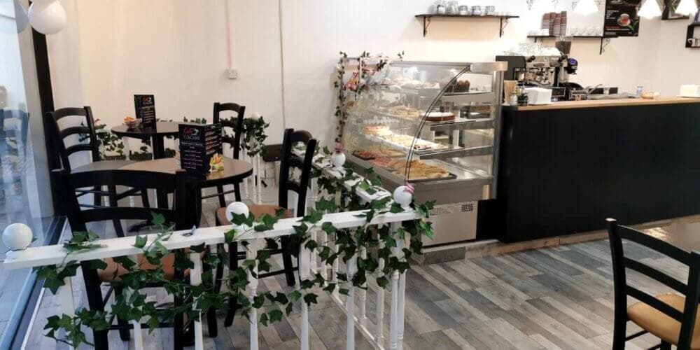 A cozy café with black chairs, small tables, and a glass display of pastries. Green vines decorate the space. - Home Instead