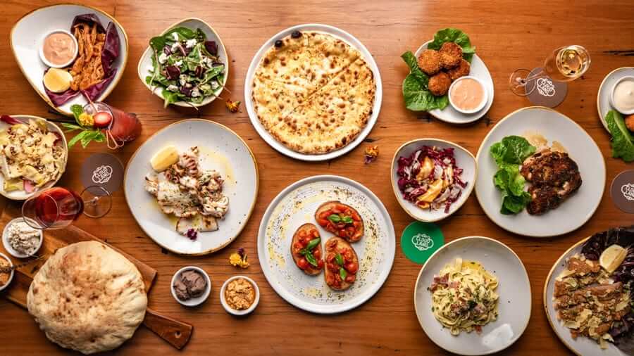 A variety of Italian dishes, including pasta, pizza, and salad, are arranged on a wooden table with drinks. - Home Instead