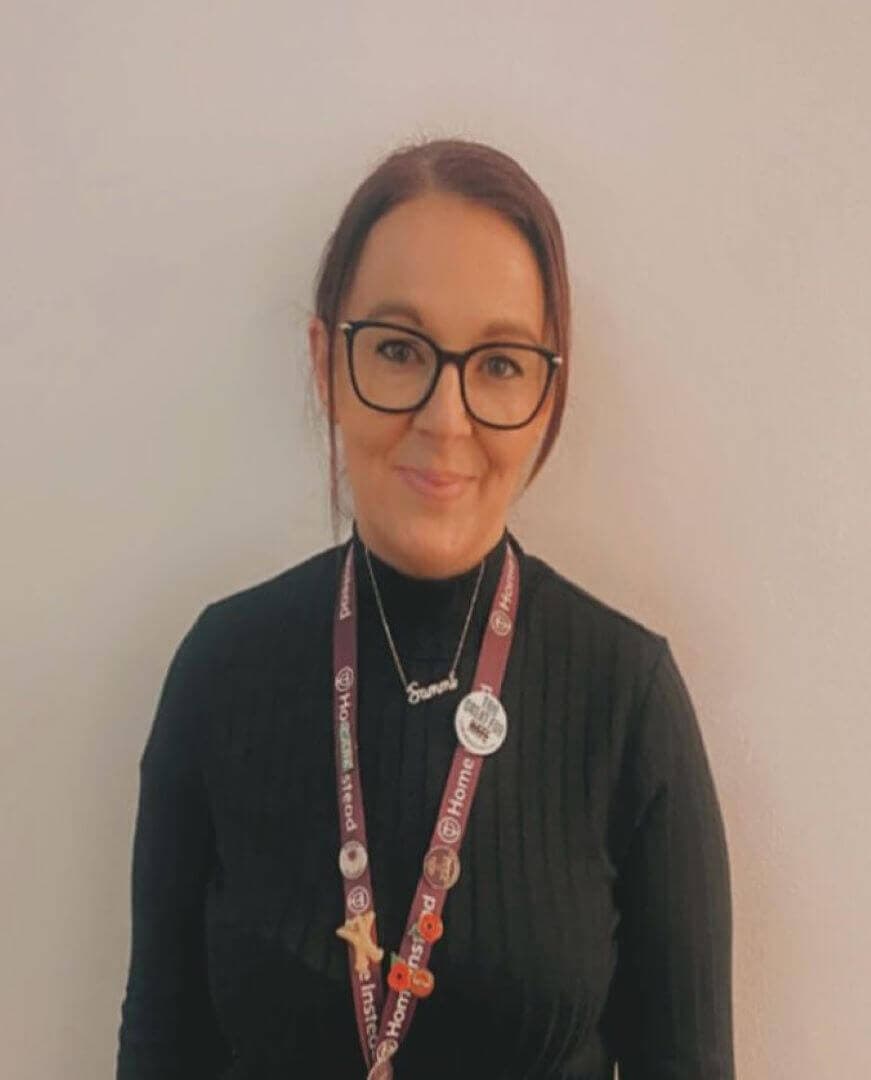 Person with glasses, wearing a black shirt and lanyard with various badges, smiling against a plain background. - Home Instead