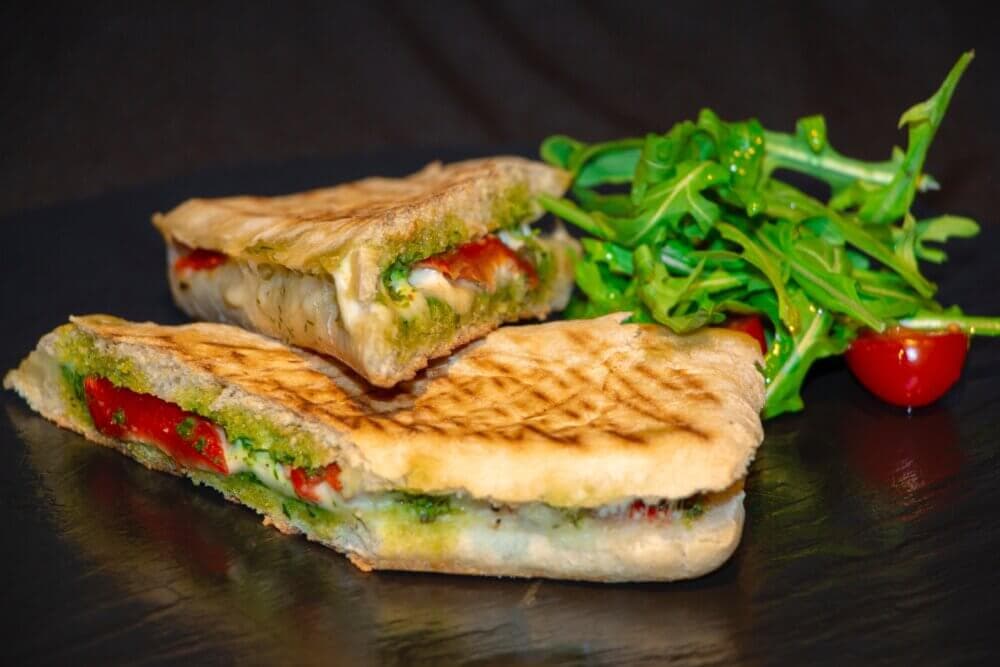 Grilled panini with melted cheese, tomatoes, and pesto, served with a side of arugula and cherry tomatoes. - Home Instead