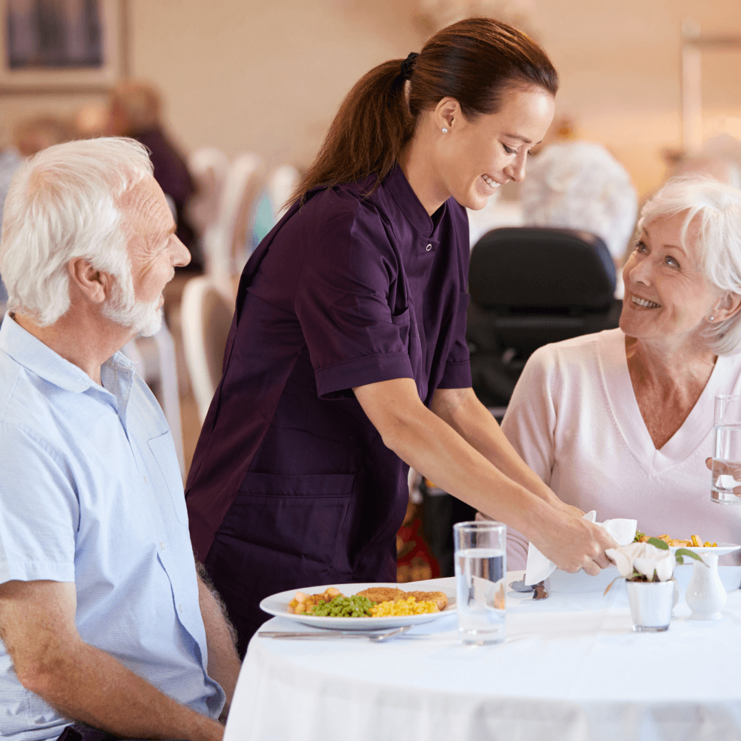 A caregiver serves a meal to an elderly man and woman seated at a dining table in a communal setting. - Home Instead