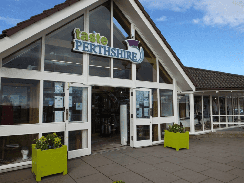 Entrance of the Taste Perthshire shop with large windows, green planters, and a "Taste Perthshire" sign above the door. - Home Instead