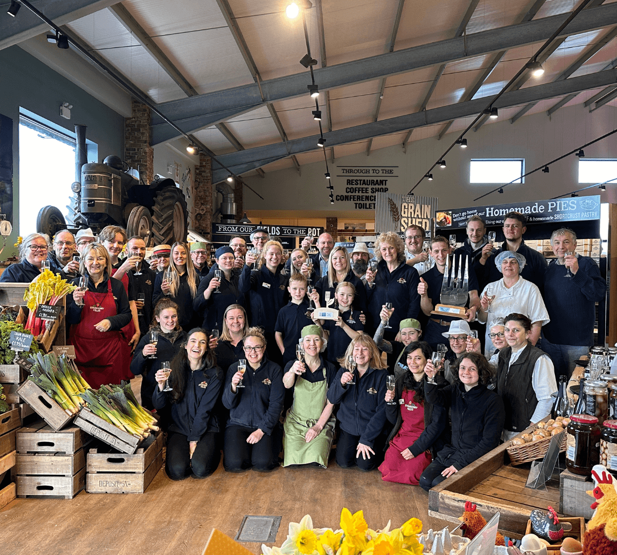 A large group of people in a market-like setting, smiling and holding glasses. Some are wearing aprons. - Home Instead