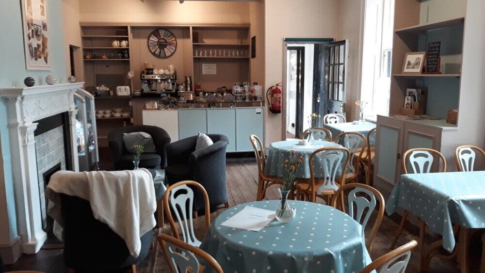 A cozy café with blue tablecloths, wooden chairs, a fireplace, armchairs, and a counter with a coffee machine. - Home Instead