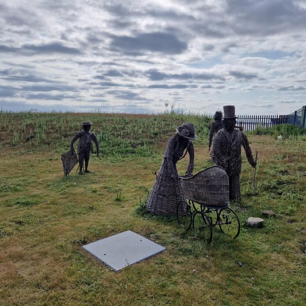 Outdoor wicker sculptures of three adults and a stroller, set in a grassy field with a cloudy sky above. - Home Instead