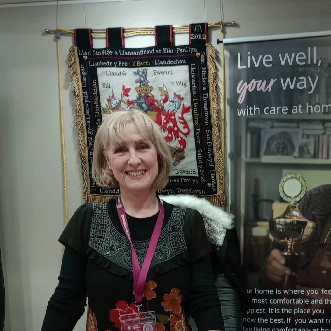 Smiling woman with blonde hair stands in front of a tapestry and a banner with health care information. - Home Instead
