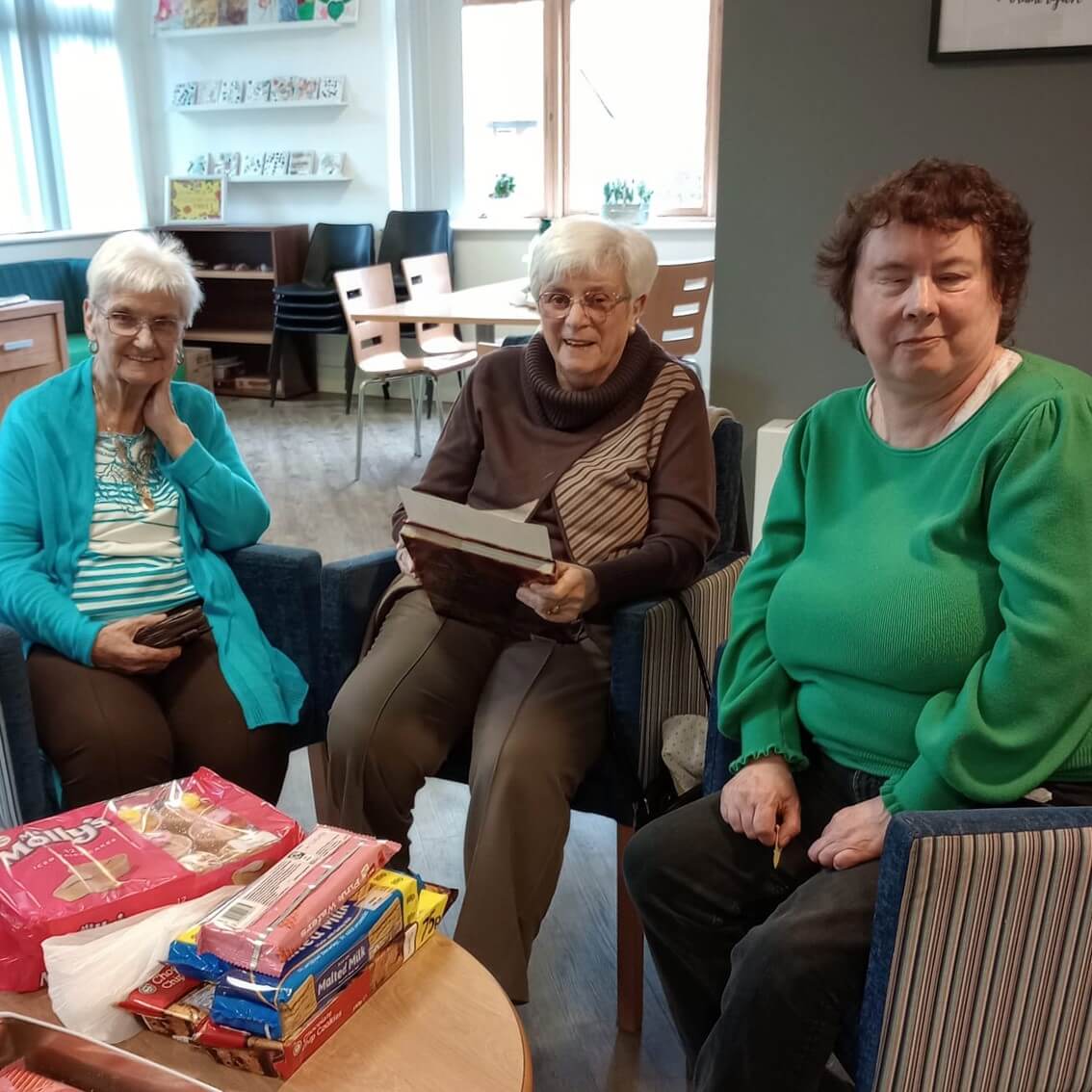 Three elderly women sitting together in a room, one holding a book, with snacks on the table in front of them. - Home Instead
