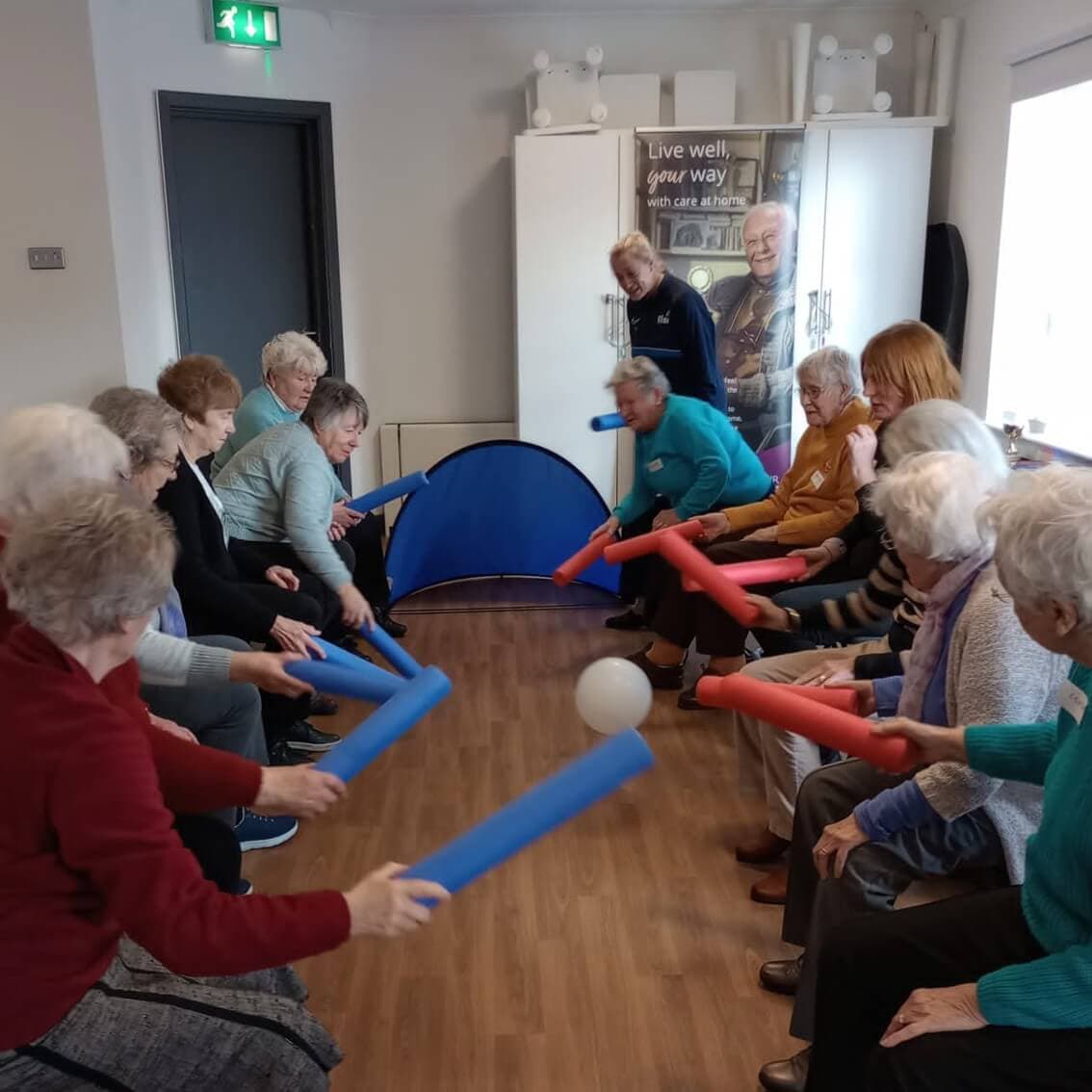 Group of elderly people sitting in a circle and playing a balloon game with pool noodles in a room. - Home Instead