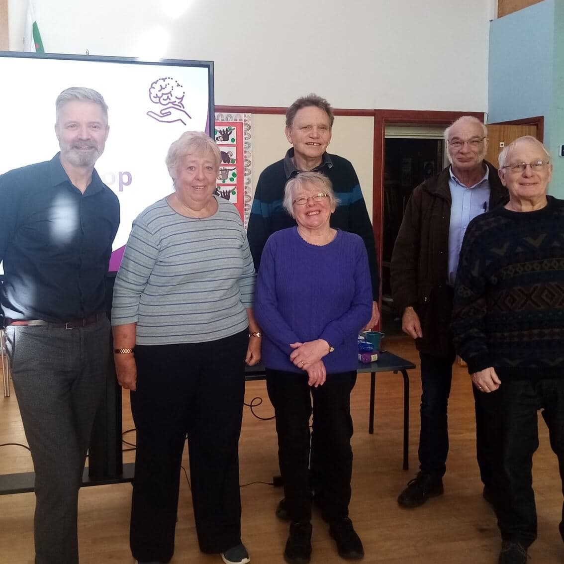 Six smiling people stand indoors with a presentation screen behind them in a well-lit room. - Home Instead