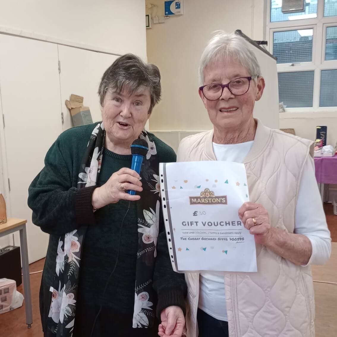 Two elderly women smiling, one holding a microphone and the other holding a £50 Marston's gift voucher in a room. - Home Instead
