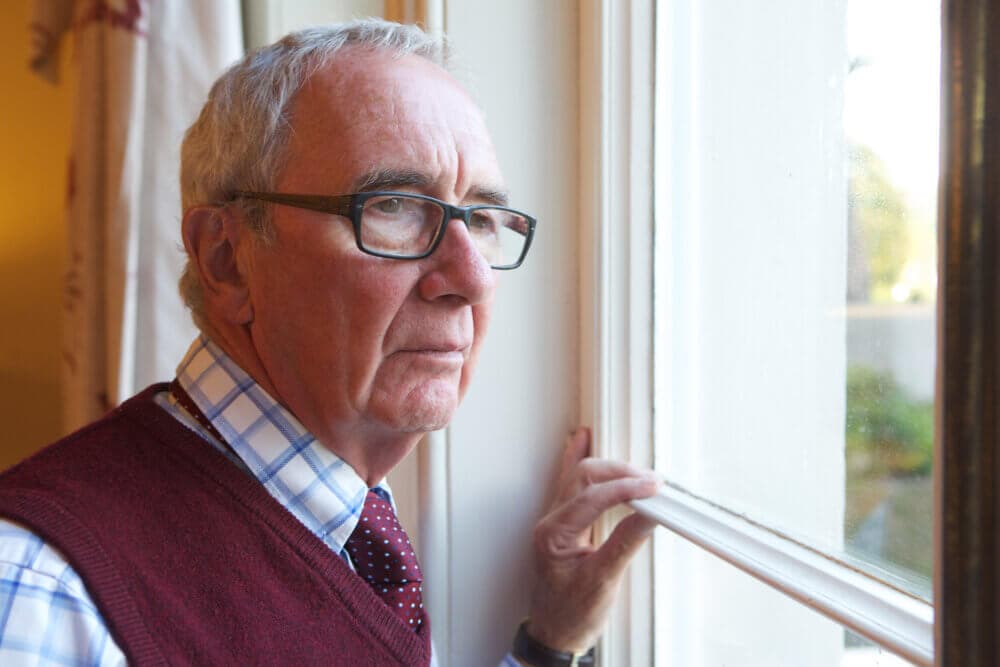 Elderly man with glasses and a red vest looking thoughtfully out a window. - Home Instead