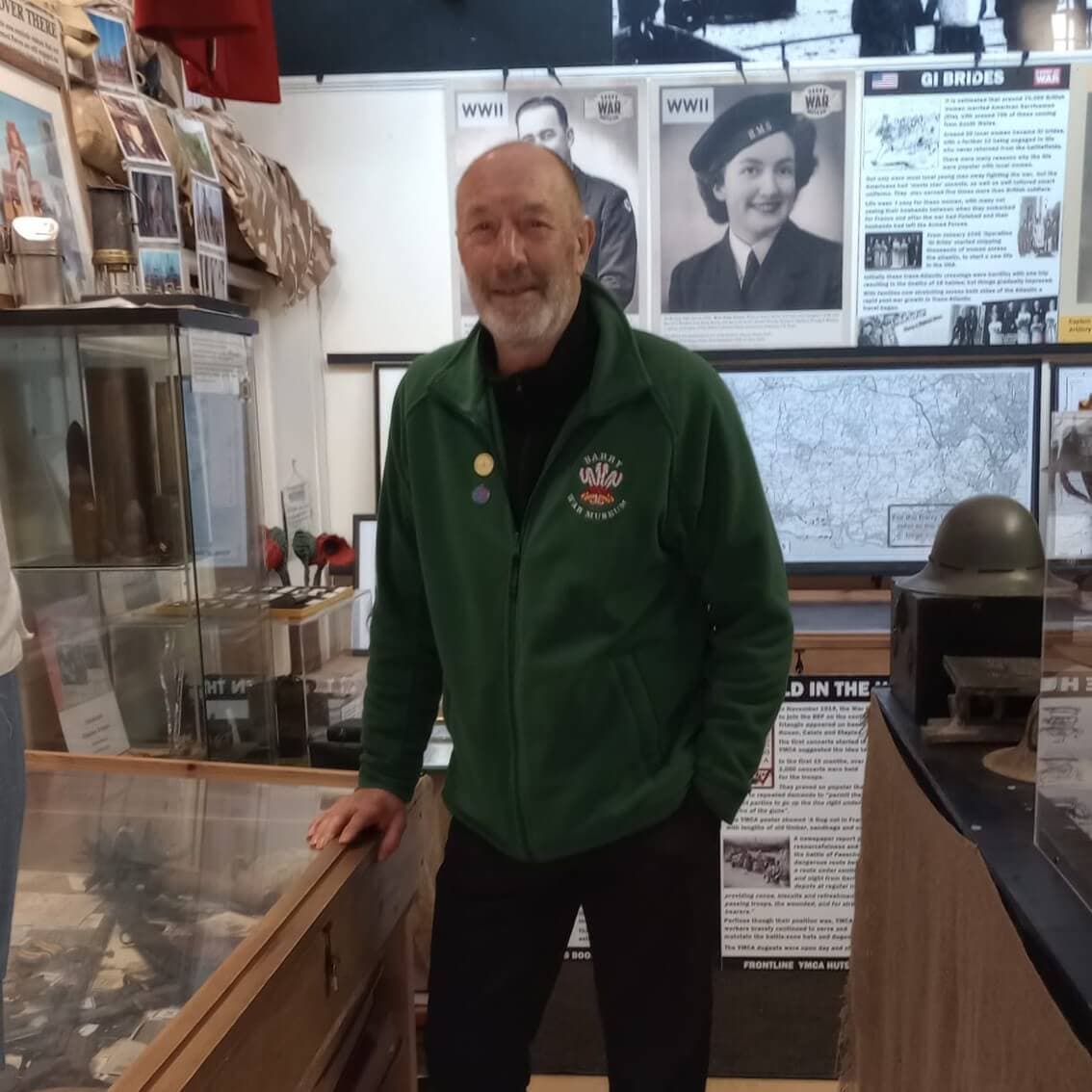 Man in a green jacket standing in a museum exhibit with WWII-era displays and historical photos on the walls. - Home Instead